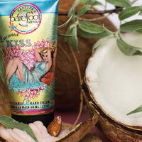 A tube of Barefoot Venus Coconut Kiss Hand Cream is placed among coconut halves and green leaves. The tube features a retro-style illustration of a woman in a blue dress, surrounded by pink and yellow floral designs. The product is 50 mL (1.7 fl oz) and boasts natural ingredients.