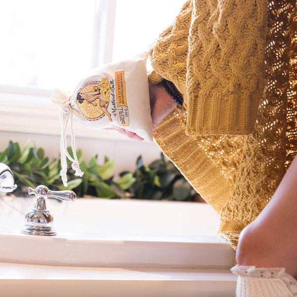 A person in a textured yellow sweater and white socks holds a Barefoot Venus 100% Natural Mustard Bath Soak Refill Bag near a bathtub, ready for a relaxing soak, with greenery and sunlight in the background.