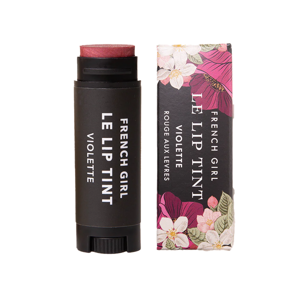 A tube of FRENCH GIRL Tinted Lip Balm in "Violette" stands beside its floral box. The sleek black balm contrasts with the packaging, which features pink and white flowers with green leaves.