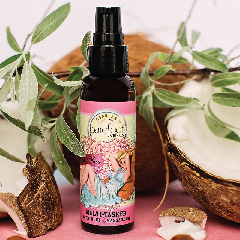 A bottle of Barefoot Venus Coconut Kiss Multi-Tasker Body+Bath+Massage Oil - Mini is displayed with leaves and coconut halves in the background, evoking a sense of island coconut. The label shows artistic imagery of a fairy and floral designs. The bottle is dark with a black pump and pink label featuring the product name and details.