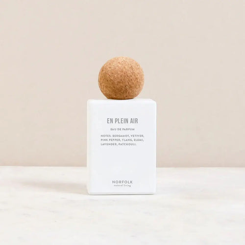 A white rectangular bottle of Norfolk Natural Living En Plein Air Parfum (50ml) with a round cork cap rests on a light surface against a beige background. The label notes bergamot and jasmine, and features the brand name at the bottom.