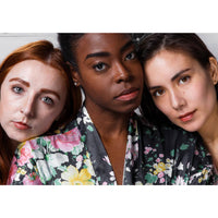 Three women with different skin tones pose together in floral outfits, showcasing natural, glowing skin and lips enhanced by FRENCH GIRL Tinted Lip Balm | Veronique. A blurred background draws attention to their radiant faces.
