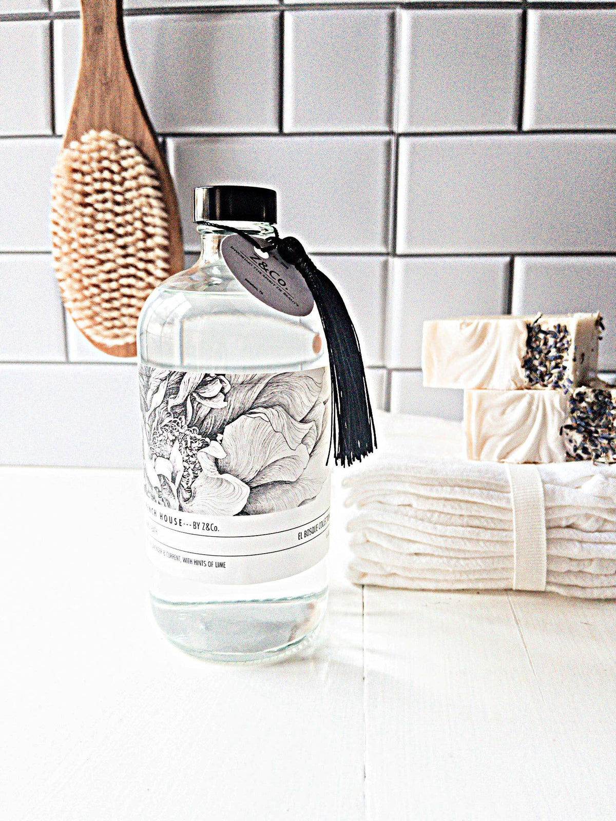 A clear glass bottle with a black cap and tassel sits on a white countertop. The Z&Co. French House El Bosque Bubble Bath label features an intricate floral design. Behind it, there are a wooden brush, two handmade soap bars with dried flowers, and a stack of white washcloths.