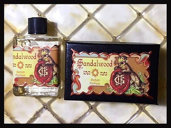 A bottle of Seventh Muse Fragrant Oil - Sandalwood, reminiscent of a French perfume, stands next to its ornate box adorned with a vintage label and decorative emblem against a textured, cream-colored surface with a diamond pattern.