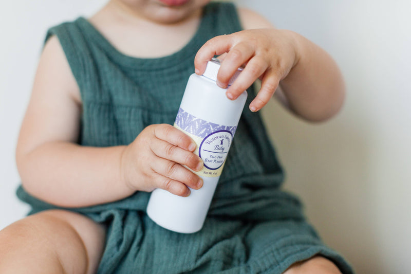 A baby in a green sleeveless outfit is holding a white bottle labeled "Victoria's Lavender Baby Powder Talc-free." The baby is focused on removing the cap of the bottle, which features chamomile and lavender. The background is out of focus, highlighting the baby and the product from Victoria's Lavender.