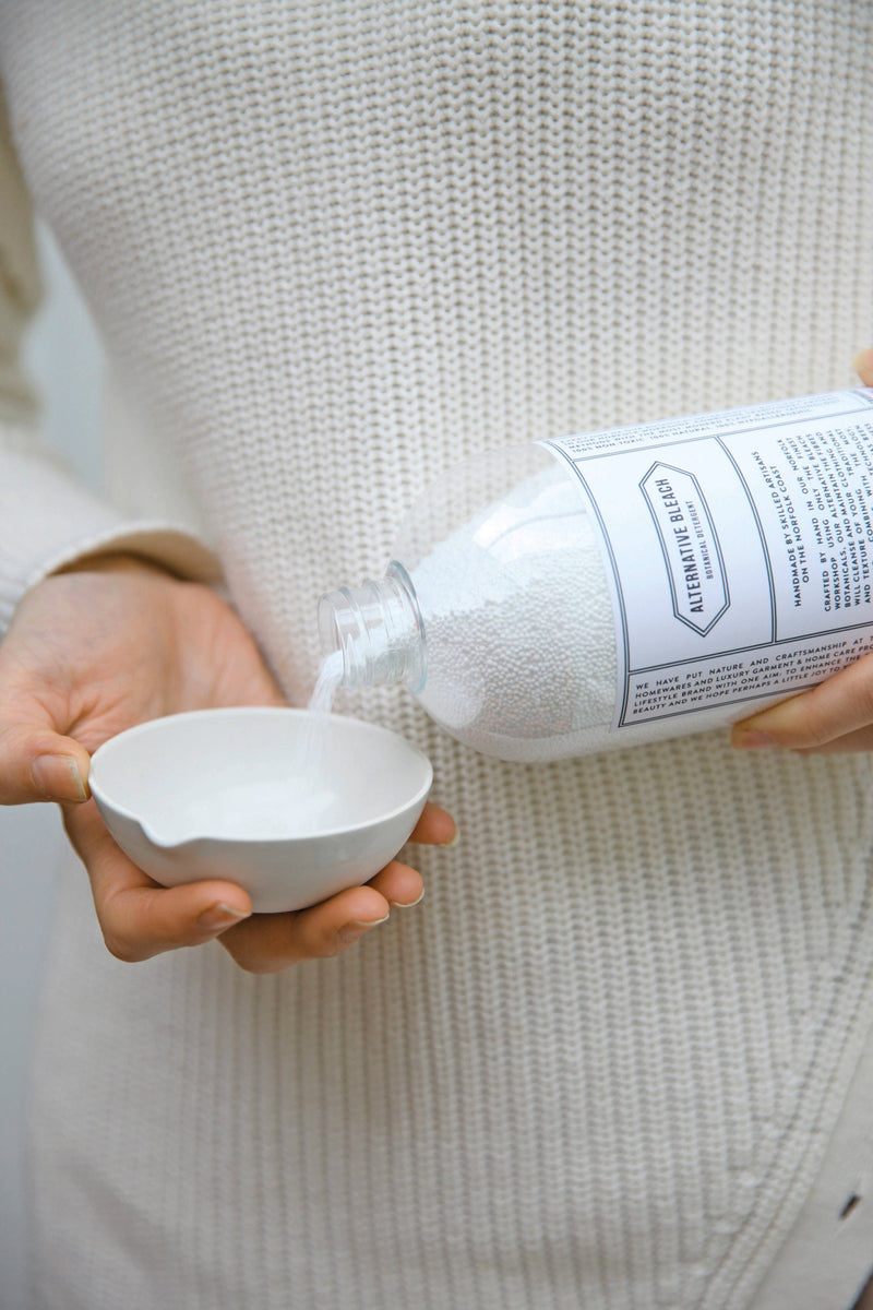A person in a white knitted sweater pours Norfolk Natural Living Alternative Bleach from a labeled bottle into a small white dish, with focus on their hands and the Norfolk Natural Living container.