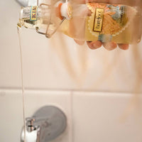 A hand holding a glass bottle labeled "Barefoot Venus" and pouring its contents—infused with natural nourishing oils—into a bathtub. The bottle, which is Barefoot Venus Coconut Kiss Body + Bath + Massage Oil, boasts a colorful design, while the faucet of the bathtub is visible in the background.