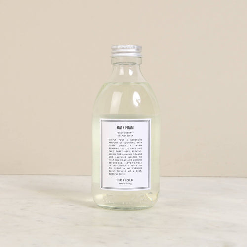 Norfolk Natural Living Deepest Sleep Bath Foam (250ml), a clear glass bottle with a silver cap, filled with transparent essential oil bath foam, rests on light marble against a beige backdrop.