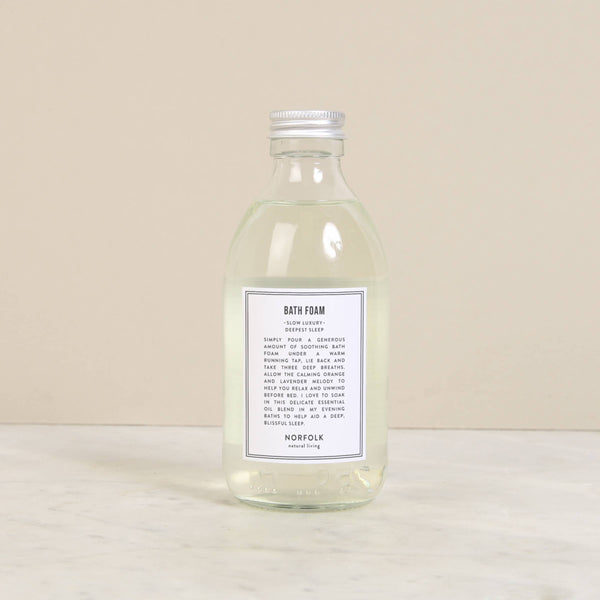 Norfolk Natural Living Deepest Sleep Bath Foam (250ml), a clear glass bottle with a silver cap, filled with transparent essential oil bath foam, rests on light marble against a beige backdrop.