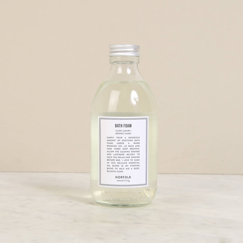Norfolk Natural Living Deepest Sleep Bath Foam (250ml), a clear glass bottle with a silver cap, filled with transparent essential oil bath foam, rests on light marble against a beige backdrop.