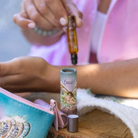 A person applies Barefoot Venus The Vanilla Effect Roll On Perfume from a roller bottle onto their wrist. Nearby, a lip balm with a fairy design and a colorful pouch rest on the textured fabric table. Wearing a pink shirt, the person enjoys the subtle allure of cruelty-free fragrance enveloping the air.