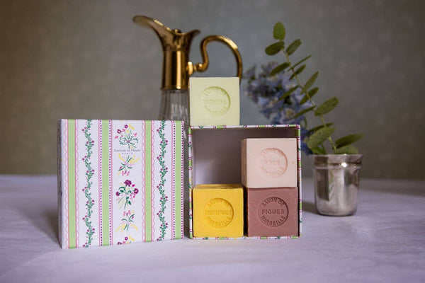 The Senteurs De France Coffret Rose Lily of the Valley, Verbena, Honeysuckle features four pastel French artisanal soaps in a decorative box, shown beside a gold pitcher, green foliage, and a silver cup on a white tablecloth.