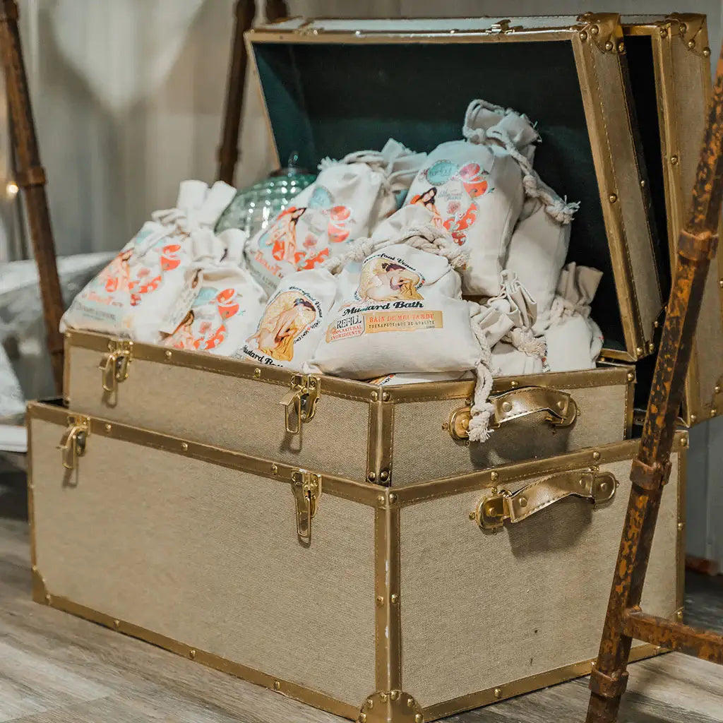 An open vintage trunk filled with colorful cloth bags, each holding a Barefoot Venus 100% Natural Mustard Bath Soak Refill Bag, sits on a wooden floor next to wooden poles.