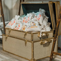 An open vintage trunk filled with colorful cloth bags, each holding a Barefoot Venus 100% Natural Mustard Bath Soak Refill Bag, sits on a wooden floor next to wooden poles.