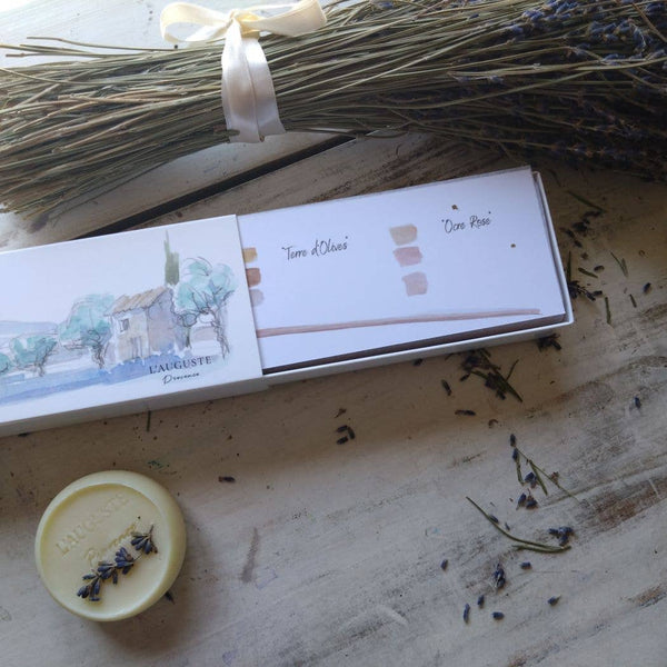 A bundle of dried lavender sits on a wooden surface beside an open watercolor box with "Terre d'Olivier" and "Ocre Rose" swatches. Nearby, a round soap with lavender sprigs exudes the essence of L'AUGUSTE Provence Handcrafted French soaps, enriched with organic vegetable oils from Provence.