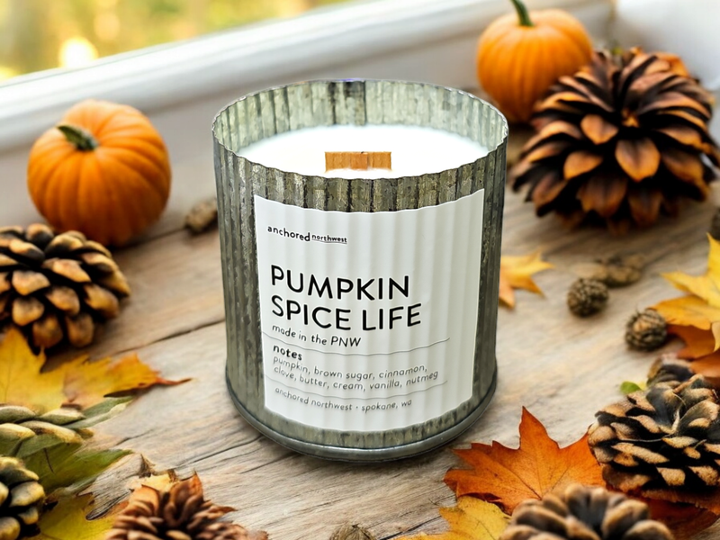 An Anchored Northwest Pumpkin Spice Life Rustic Vintage Farmhouse Wood Wick Candle glows on a wooden table, surrounded by small pumpkins, pine cones, and autumn leaves. Crafted from soy wax, it features fragrant notes of pumpkin, brown sugar, cinnamon, clove, butter, cream, vanilla, and nutmeg.