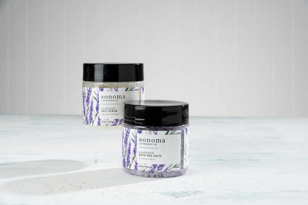 Two jars of Sonoma Lavender Sea Salt Scrub by Sonoma Lavender are shown on a light surface with a textured white background. Infused with calming lavender essential oil, they gently exfoliate and moisturize your skin.