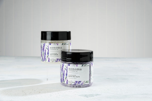 Two jars of Sonoma Lavender Sea Salt Scrub by Sonoma Lavender are shown on a light surface with a textured white background. Infused with calming lavender essential oil, they gently exfoliate and moisturize your skin.