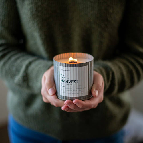 A person wearing a green sweater is holding a lit candle in a ribbed, cylindrical container labeled "Fall Harvest." Made with 100% American-grown soy wax, this Anchored Northwest Fall Harvest Rustic Vintage Farmhouse Wood Wick Candle adds charm to any home decor. The background is softly blurred, focusing attention on the candle and the person's hands.