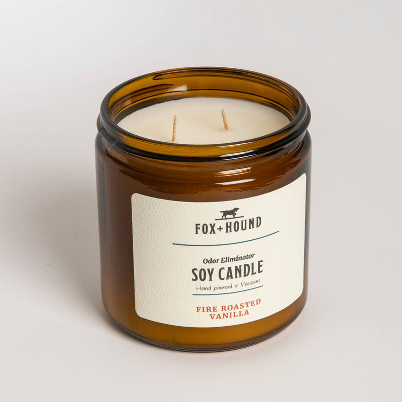 A glass jar soy candle with a beige label, featuring the text "Fox + Hound, odor-eliminating soy candles, fire roasted vanilla, hand poured in Wisconsin" on a plain white background.