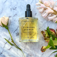 A 2 oz bottle of FRENCH GIRL Organic Hydrating Jasmine Body Oil with a dropper rests on marble, surrounded by soft pink and green flowers.