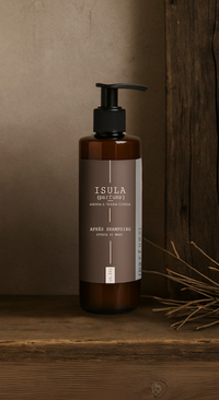 A brown pump bottle labeled “Isula Storia di Mare - Sea Story Hair Conditioner” by ISULA parfums sits on wood beside a rustic panel and dried grass, evoking a natural, earthy Mediterranean vibe.