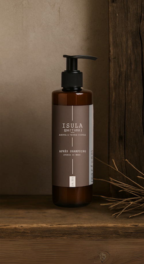 A brown pump bottle labeled “Isula Storia di Mare - Sea Story Hair Conditioner” by ISULA parfums sits on wood beside a rustic panel and dried grass, evoking a natural, earthy Mediterranean vibe.