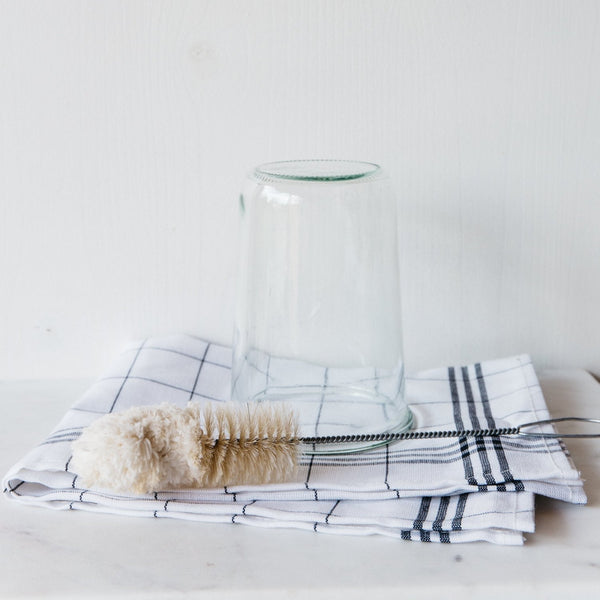 An Andrée Jardin Bistro Flute Cleaning Brush with a metal handle and cream bristles sits before a clear glass on white grid-patterned dish towels, ready to clean champagne flutes—an ideal barware maintenance setup.