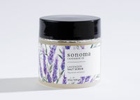 A 22 oz (623 g) Sonoma Lavender Sea Salt Scrub jar with a black lid, purple lavender designs, and text promising to "rejuvenate and restore" as you exfoliate and moisturize your skin. Infused with lavender essential oil.