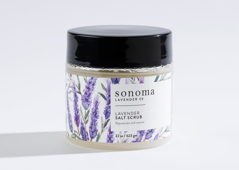 A 22 oz (623 g) Sonoma Lavender Sea Salt Scrub jar with a black lid, purple lavender designs, and text promising to "rejuvenate and restore" as you exfoliate and moisturize your skin. Infused with lavender essential oil.