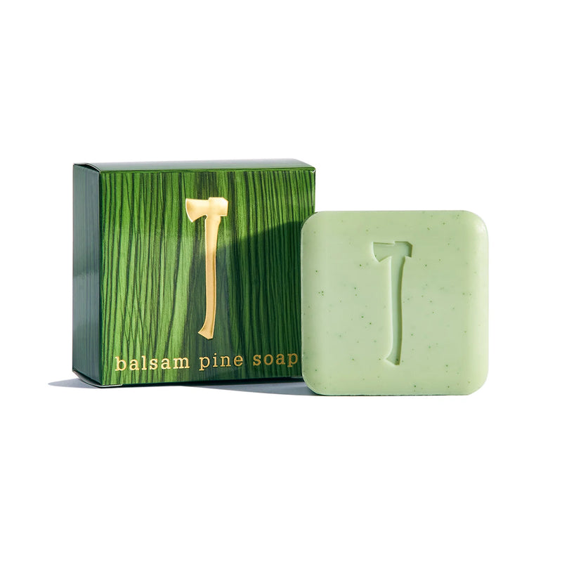 The aromatic scents of balsam pine fill the air as a green bar of Kala's Balsam Pine Bar Soap rests beside its matching box, both embossed with an axe design. The packaging, featuring a wood grain pattern, complements the soap's natural exfoliators for an invigorating experience.