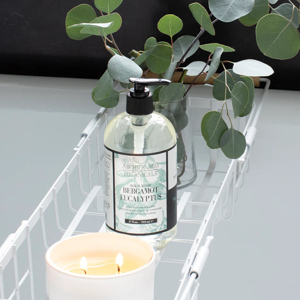 A bottle of Archipelago Botanicals Bergamot Eucalyptus Body Wash 17oz rests on a white metal rack beside a lit white candle and fresh eucalyptus branches.