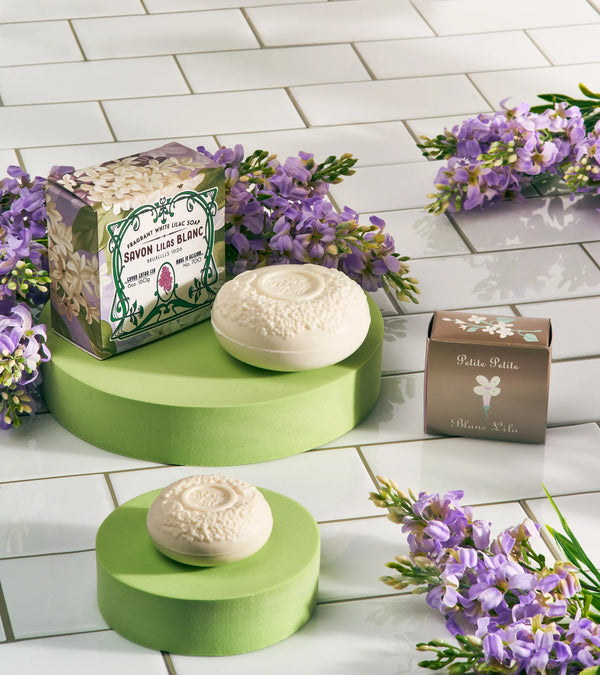 A decorative display showcases two round Blanc Lila Lilac Bath Soaps from the Soaps, Soaks & Pampering Products From Around the World brand. One soap is partially unwrapped, revealing ornate patterns, and both are placed on green pedestals. Accompanying the soaps are two boxes—one floral and one plain—surrounded by purple flowers, all set on a white tiled surface. The arrangement exudes an intoxicating fragrance of lilac.