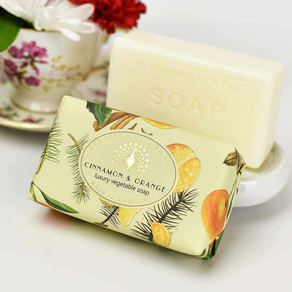 Two bars of The English Soap Co. Cinnamon Orange Vintage Wrapped Soap, one wrapped in a cinnamon and orange themed paper, set against a backdrop of a floral teacup and fresh flowers.