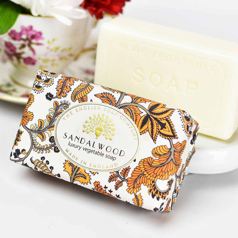 A bar of The English Soap Co. Sandalwood Vintage Italian Wrapped Soap with ornate orange and black packaging, next to another bar of vegetable soap and a vase with red flowers.