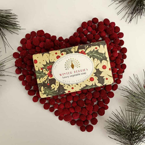 A bar of The English Soap Co. Winter Berries Festive Christmas Soap, part of our Christmas Collection, is displayed on a heart-shaped arrangement of red berries. The vegan-friendly soap packaging features a festive design with holly leaves and berries, while pine branches frame the image's corners against a white background.