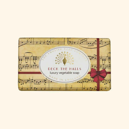 The English Soap Co. Deck The Halls Christmas Soap is a luxury, vegan-friendly vegetable soap that features shea butter for a rich lather. Its packaging showcases musical notes with a decorative red ribbon and bow, along with an oval label in the center adorned with gold accents. Made in England by The English Soap Co.