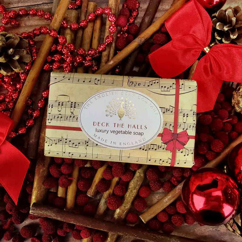 A rectangular bar of The English Soap Co. Deck The Halls Christmas Soap, featuring a music sheet design with the text "Deck the Halls, luxury vegetable soap, Made in England," is surrounded by cinnamon sticks, pinecones, red berries, red bows, red beads, and red Christmas ornaments. This vegan-friendly soap from The English Soap Co. also contains shea butter.