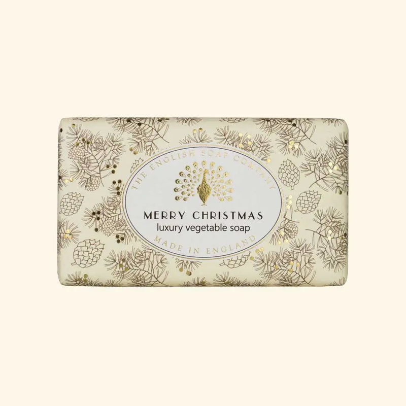 A cream-colored, rectangular bar of The English Soap Co. Merry Christmas Soap is wrapped in festive paper adorned with gold and brown pinecones and foliage. The label reads "The English Soap Company" and "Merry Christmas luxury vegetable soap made in England with shea butter, vegan friendly.