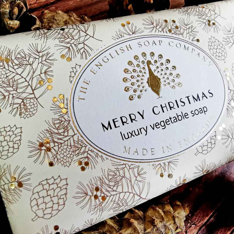 An elegant The English Soap Co. Merry Christmas Soap box from The English Soap Company. The packaging features a festive pinecone design with golden accents, a central label with a peacock illustration, and text reading, "Merry Christmas luxury vegetable soap with shea butter, Made in England." Vegan friendly.