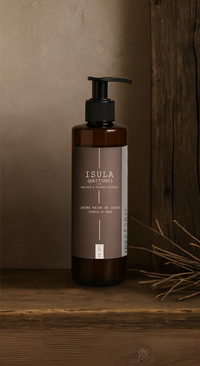 A brown pump bottle labeled "ISULA parfums" sits on rustic wood beside dried grass, showcasing ISULA Storia di Mare - Sea Story Hand and Body Cream—a hydrating moisturizer with a refreshing marine scent.