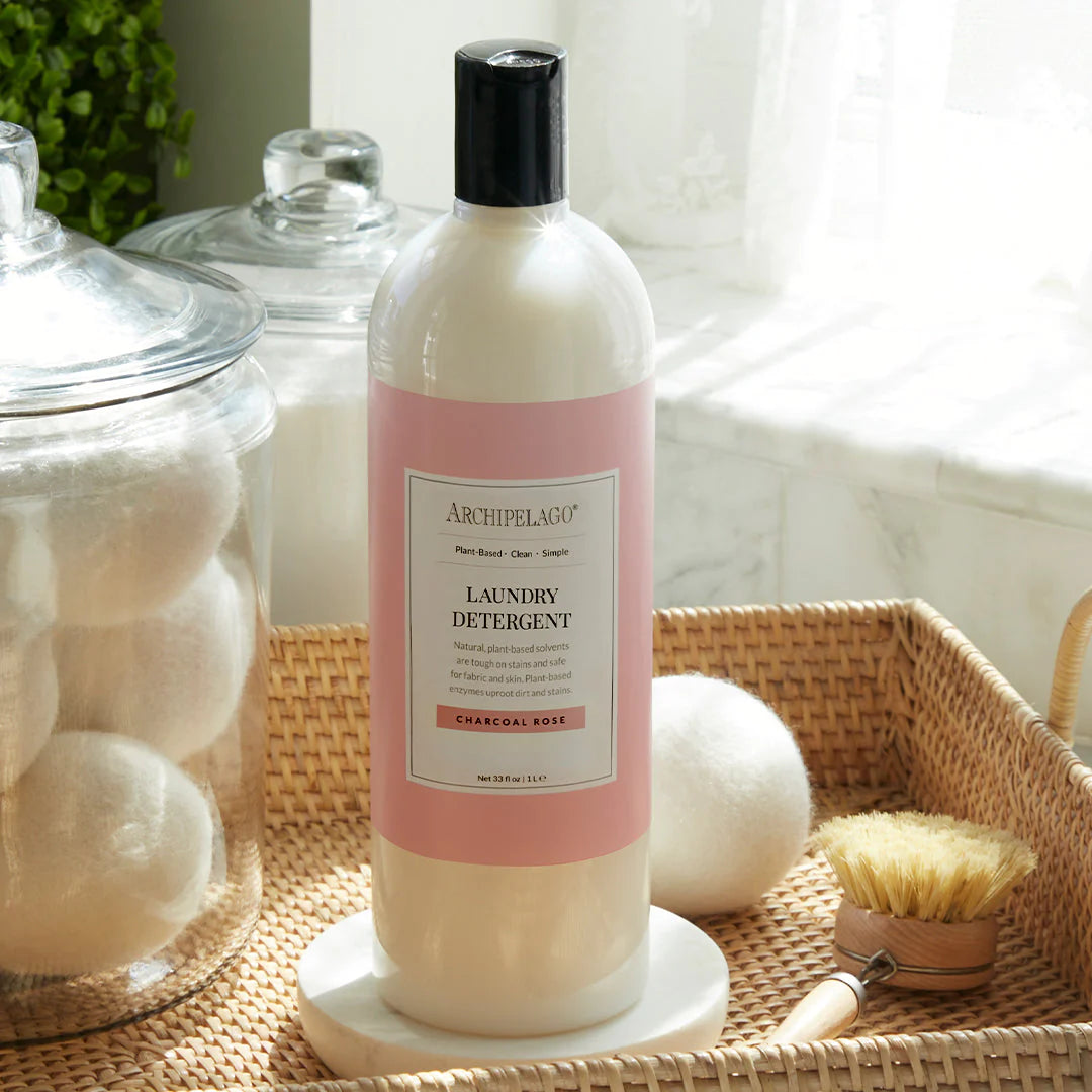 A bottle of Archipelago Charcoal Rose Laundry Detergent, featuring bio-based ingredients, sits on a white tray beside a brush, with jars of wool dryer balls in the background, illuminated by natural light.