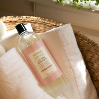 A bottle of Archipelago Charcoal Rose Laundry Detergent rests on a fluffy white towel inside a woven basket, illuminated by warm, natural sunlight.