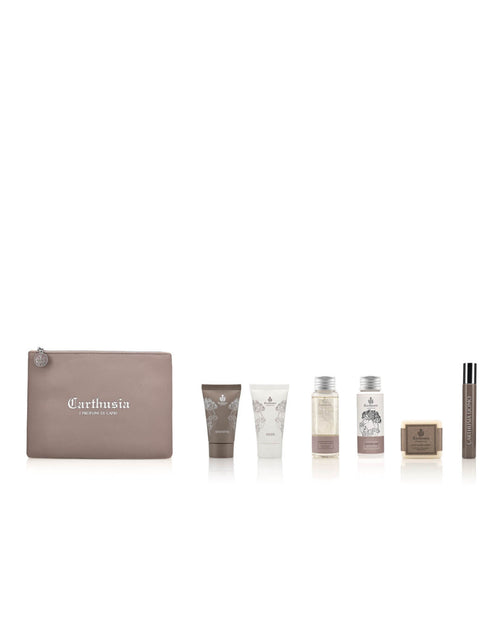 The image shows a set of travel-sized Carthusia Uomo personal care products. It includes a taupe zippered Carthusia Uomo Travel Clutch Bag, two tubes, two small bottles, a bar of soap, and a slim cylindrical tube, all arranged in a row against a plain white background. Perfectly curated as part of the Men's Line by Carthusia I Profumi de Capri.