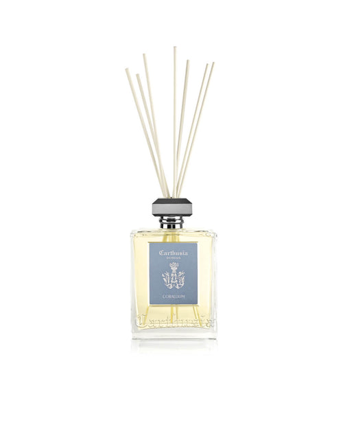The Carthusia I Profumi di Capri Corallium Reed Diffuser (230ml) features clear glass, light yellow liquid, a gray cap, pale blue label, and reed sticks for a woody aromatic home fragrance, shown against a white background.