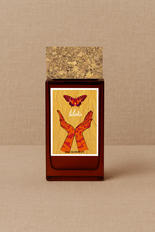 ISULA parfums LIberté eaux de parfum by ISULA parfums features a cork-capped bottle with a yellow label showing two hands with orange patterns reaching for a red butterfly, evoking a Corsica-inspired fragrance. The bottle rests on a beige surface.