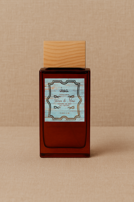 A rectangular amber glass bottle of ISULA parfums Storia di Mare - Sea Story, topped with a light wooden cap, rests on a beige textured surface. Its vintage blue and white label evokes an aquatic scent with decorative borders and cursive text.