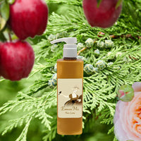 A pump bottle of La Bouquetiere Embrasse Moi Body Lotion featuring a vintage-style label stands before greenery, with apples, juniper berries, and a peach-pink rose in the background.