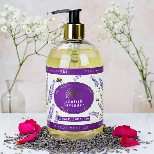 A bottle of The English Soap Co. English Lavender Hand Wash, enriched with moisturizing liquid soap, is surrounded by dried lavender buds and scattered rose petals, with a backdrop of delicate baby's breath flowers. The clear bottle, featuring eco-friendly packaging and a purple label adorned with lavender illustrations, holds 500ml of the exquisite hand wash.
