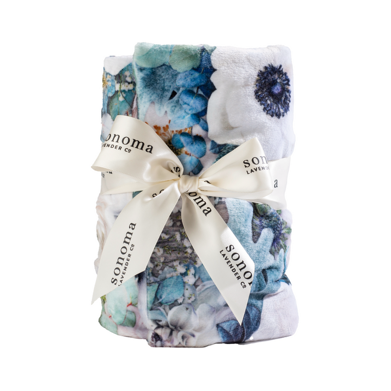 A Sonoma Lavender Eucalyptus Blooming Buds Heat Wrap with hues of blue and white, neatly folded and secured with an elegant cream ribbon that has the word "Sonoma" printed on it.